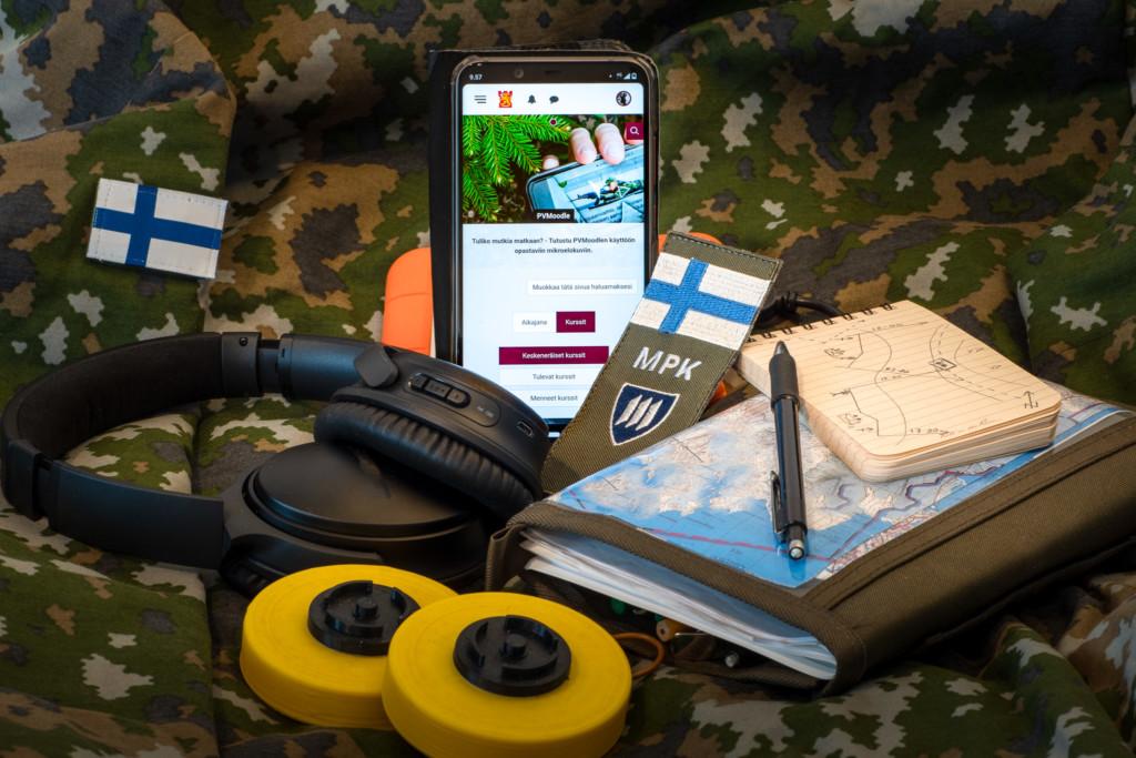 Kännykkä, kuulokkeet, muistiinpanovälineet ja MPK:n hihamerkki maastokuvioisella taustalla. Kännykässä auki PVMoodlen etusivu.