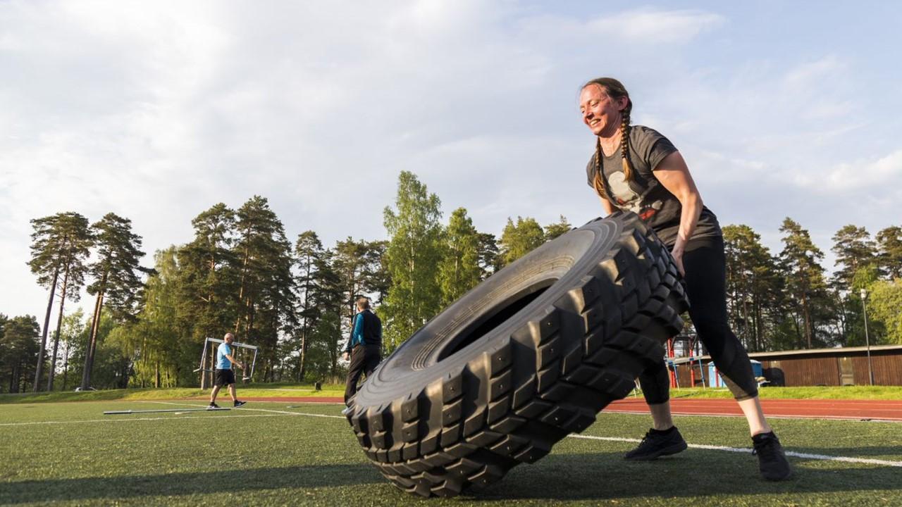 Nainen nostaa maastokuorma-auton rengasta Military Cross Training -harjoituksessa urheilukentällä.