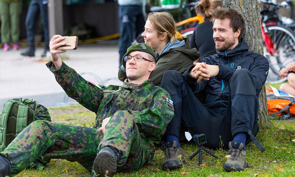 Maastopukuinen reserviläinen sekä siviiliasuinen nuori nainen ja mies istuvat yhdessä puun juurella ottamassa valokuvaa puhelimella.