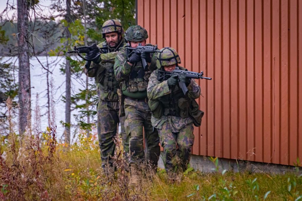 Kolme sotilasta etenee tiiviissä muodostelmassa punaisen talon nurkalla.