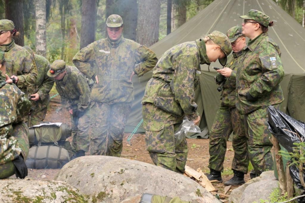 Naisia ulkona maastopuvussa nuotion äärellä