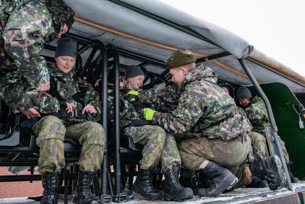 Nuoria naisia istumassa Puolustusvoimien henkilökuljetuskaluston penkeillä, varusmies opastamassa.