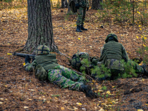 Kaksi maastopukuista henkilöä harjoittelemassa taisteluensiapua. Kolmas henkilö tähystää rynnäkkökiväärillä.