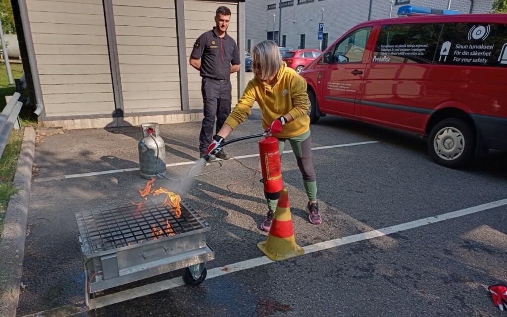 Nainen keltaisessa hupparissa sammuttaa tulta käsisammuttimella. Taustalla pelastuslaitoksen asussa oleva mies.