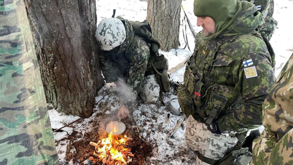 Kaksi miestä maastopuvuissa keittää vettä nuotiolla