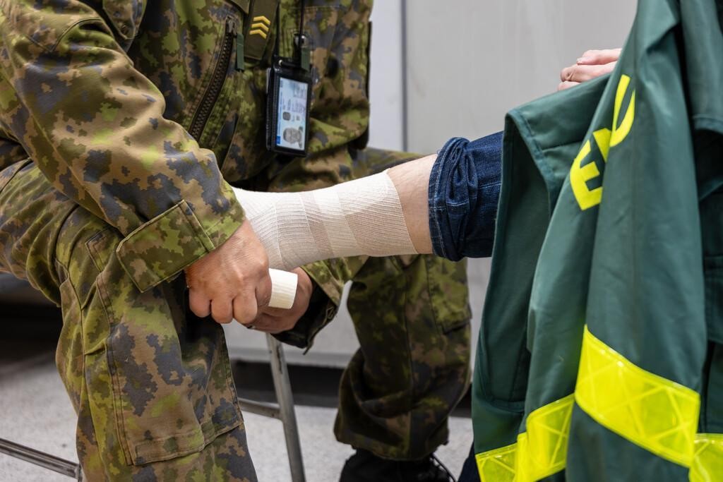 Kuvassa maastopukuun pukeutunut henkilö sitoo toisen miehen nilkkaa. 