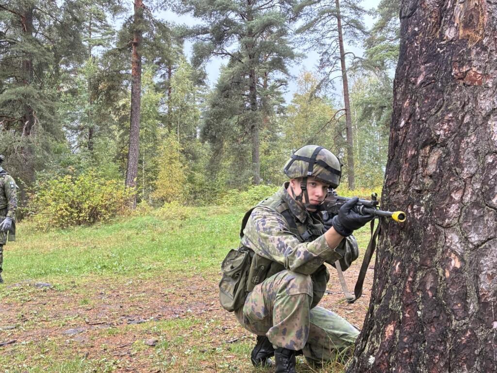 Kuvassa on mies, joka on korkeassa polviasennossa puun vieressä. Hänellä on maastopuku, kypärä ja rynnäkkökivääri.