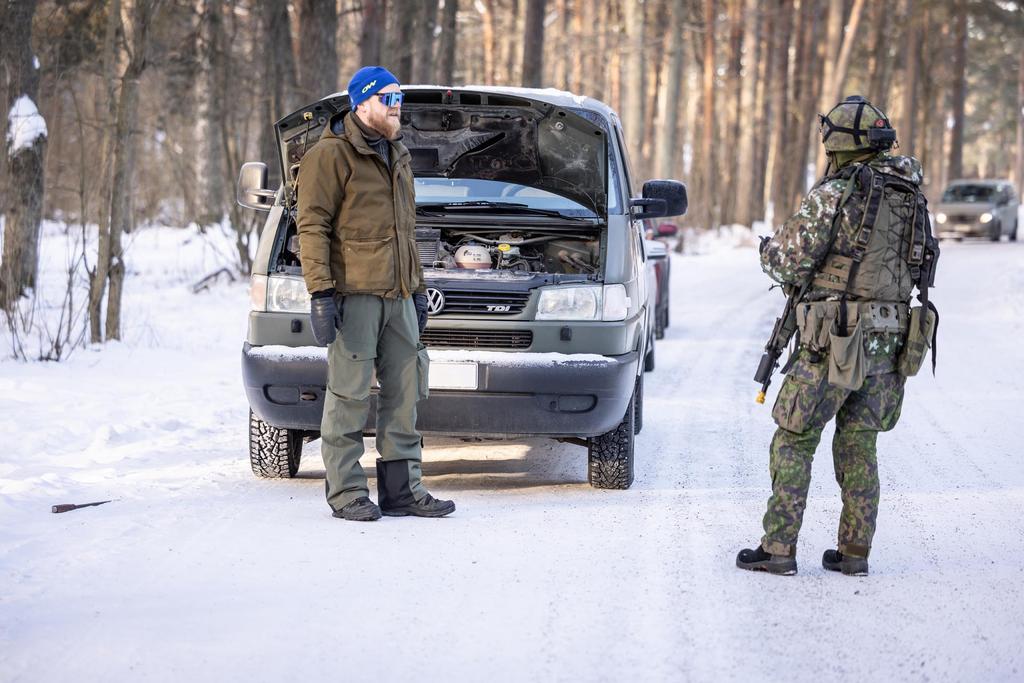 Mies seisoo pakettiauton edessä metsätiellä, sotilas tarkastaa mies-auto yhdistelmää. Muita autoja taustalla.